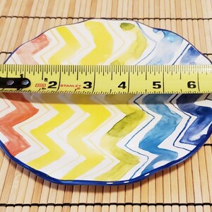 May include: A colorful, oval-shaped ceramic plate with a vibrant chevron pattern in red, yellow, green, and blue. The plate measures approximately 7 inches in length and has a dark blue border.