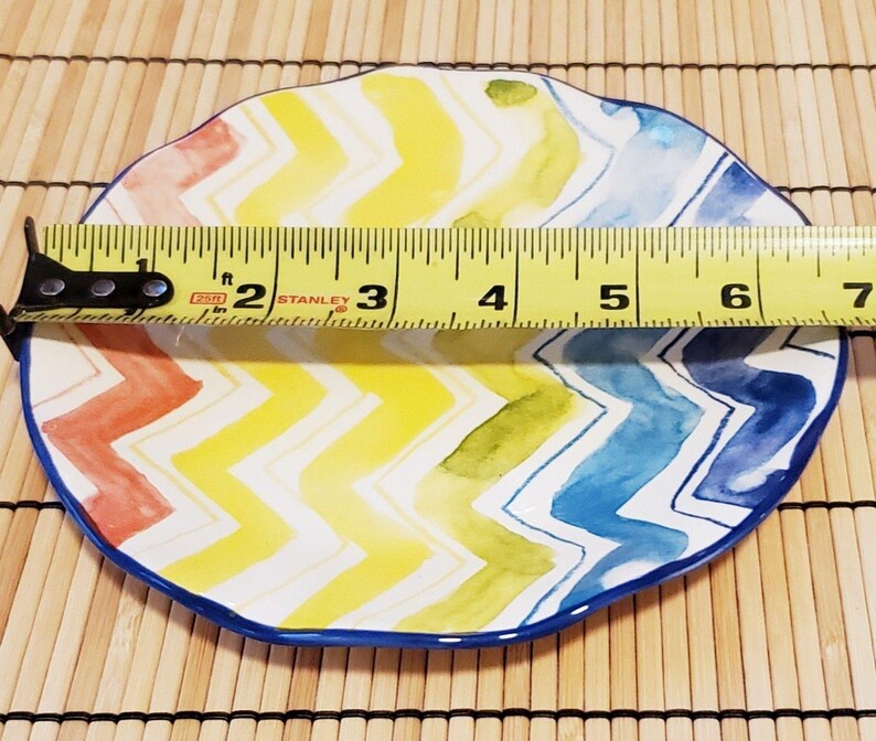 May include: A colorful, oval-shaped ceramic plate with a vibrant chevron pattern in red, yellow, green, and blue. The plate measures approximately 7 inches in length and has a dark blue border.