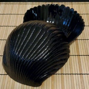 May include: A glossy black decorative seashell-shaped bowl. The bowl has a scalloped rim and a textured, ridged surface. The bowl is sitting on a bamboo mat.