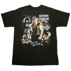 May include: Black t-shirt featuring a graphic of a wrestler with the text "STONE COLD" and "WWF". The shirt has a crew neck and short sleeves. The graphic includes images of the wrestler in action and his signature.