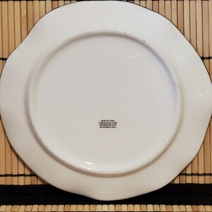 May include: A white ceramic plate with a wavy edge and a black trim. The plate has a circular center and a label that reads "Made in China, Fabriqu&eacute; en Chine, Dishwasher Safe, Microwave Safe."