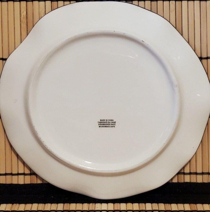 May include: A white ceramic plate with a wavy edge and a black trim. The plate has a circular center and a label that reads "Made in China, Fabriqu&eacute; en Chine, Dishwasher Safe, Microwave Safe."