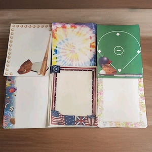 May include: A collection of decorative stationery featuring various designs. The stationery includes a baseball theme, tie-dye, a baseball field, and patriotic borders with flags. Each piece has a blank space for writing.