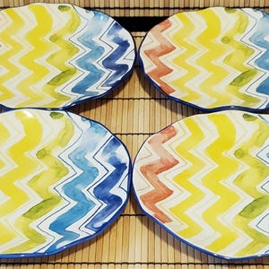 May include: Four decorative ceramic plates with a colorful zigzag pattern in yellow, blue, red, and green. Each plate has a wavy edge and a blue border. The plates are arranged on a bamboo mat.