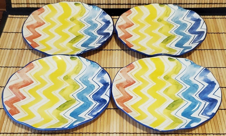 May include: Four decorative ceramic plates with a colorful zigzag pattern in yellow, blue, red, and green. Each plate has a wavy edge and a blue border. The plates are arranged on a bamboo mat.