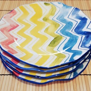 May include: A stack of four ceramic plates with a wavy edge. Each plate features a colorful zigzag pattern in shades of red, yellow, green, and blue. The plates have a blue border and are stacked on top of each other.