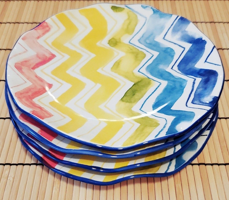 May include: A stack of four ceramic plates with a wavy edge. Each plate features a colorful zigzag pattern in shades of red, yellow, green, and blue. The plates have a blue border and are stacked on top of each other.