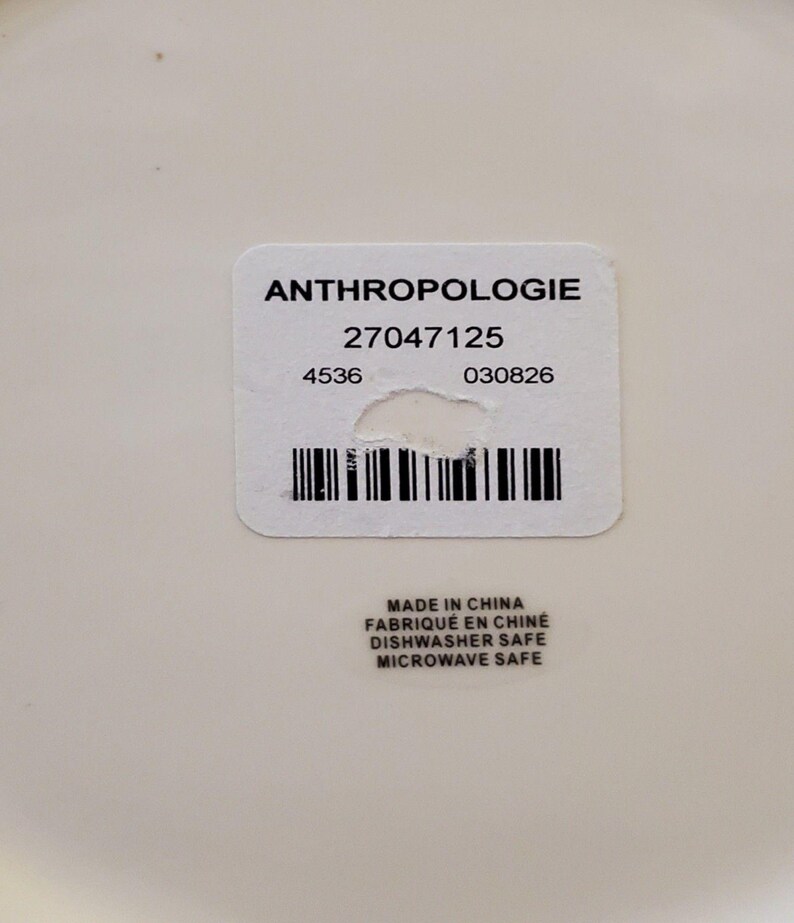 May include: A close-up of a white label with the word "ANTHROPOLOGIE" printed in black. Below the word are numbers and a barcode. The label also includes the text "MADE IN CHINA", "DISHWASHER SAFE", and "MICROWAVE SAFE".