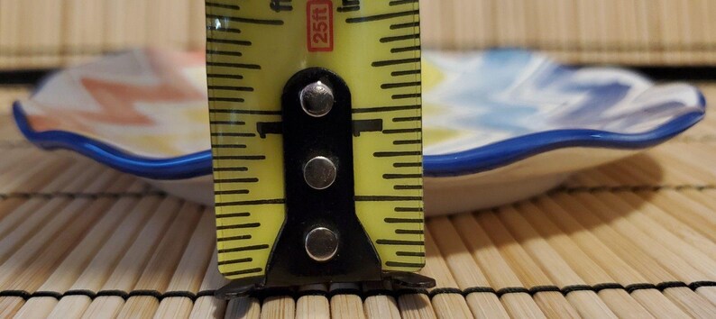 May include: A small, decorative ceramic dish with a wavy edge and a blue rim. The dish features a colorful, abstract design in shades of orange, yellow, and blue. The dish is approximately 1 inch in height.