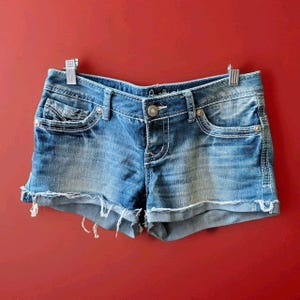 Op de afbeelding: Blauwe spijkershort met een medium wassing, een knoop- en ritssluiting, vijf zakken met zilverkleurige klinknagels en een gerafelde zoom. De tailleband is voorzien van riemlussen en witte contraststiksels over het hele kledingstuk.