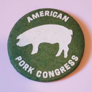 May include: A round, green item with the words "AMERICAN" and "PORK CONGRESS" in white lettering. A white silhouette of a pig is in the center. The item has a textured surface.
