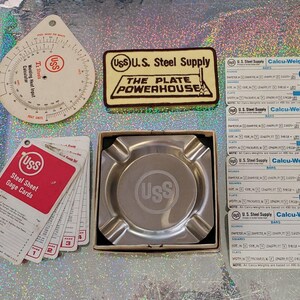 May include: A collection of U.S. Steel Supply promotional items including a circular welding heat input calculator, a rectangular sign reading The Plate Powerhouse, a set of red and white steel sheet gage cards, and a silver metal ashtray with a USS logo.