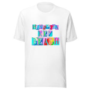 Puede incluir: Una camiseta blanca de manga corta con un estampado gráfico en el pecho. El diseño consiste en el texto MANASOTA KEY BEACH mostrado en letras coloridas, en bloque y multicolores sobre una variedad de formas rectangulares brillantes.