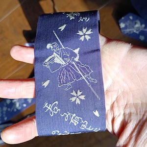 Puede incluir: Una tela azul marino con una ilustración blanca de un guerrero samurái con una espada, patrones florales y de escritura. La tela se sostiene en una mano.