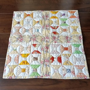 Può includere: Una trapunta fatta a mano con un motivo ripetuto di forme a farfalla colorate. La trapunta presenta uno sfondo color crema con motivi floreali ed è accentuata da quadrati di tessuti rossi, arancioni, gialli, verdi e blu.