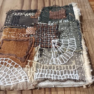 May include: Textile art piece with a patchwork design. The piece features squares of brown, black, and green fabric, with white embroidered web-like patterns. The edges are frayed, and the piece is set on a wooden surface.