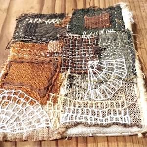 May include: A textile art piece composed of various fabric patches in shades of brown, black, and cream. The patches are stitched together, with some featuring intricate white lace-like designs. The edges are frayed, giving it a rustic, handmade appearance.