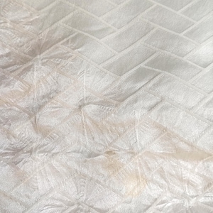 May include: Close-up of a white fabric with a textured pattern. The pattern features a repeating design of rectangular shapes arranged in a brick-like pattern, with a subtle sheen. The fabric appears soft and slightly wrinkled.