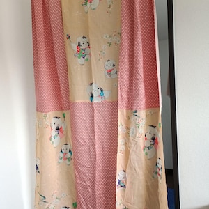 Pode incluir: Uma cortina decorativa com um design de patchwork. A cortina tem um painel central com figuras de desenhos animados e detalhes florais em um fundo de pêssego claro. Os lados são um padrão de bolinhas rosa.