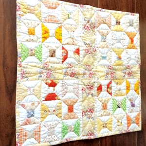Op de afbeelding: Een vierkante stoffen quilt met een herhalend klospatroon in diverse kleuren, waaronder geel, oranje, groen en rood. Het ontwerp bevat bloem- en ruitvormige stoffen lapjes gescheiden door witte driehoekige segmenten met zichtbare stiksels.