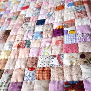 May include: A handmade patchwork quilt with a variety of fabrics in a grid pattern. The quilt features squares of different colors and patterns, including floral, polka dots, and solid colors. The color palette includes pinks, purples, blues, and creams.