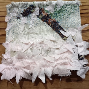 Op de afbeelding: Een klein, getextureerd textielkunstwerk met een lichtgroene en witte achtergrond. Een donker, patroonachtig element is diagonaal in het midden geplaatst. De onderste rand is versierd met lichtroze stoffen stroken.