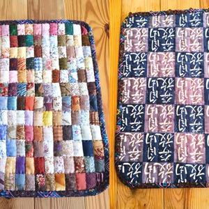 Puede incluir: Dos cojines de asiento acolchados. Uno presenta un diseño de patchwork con varios patrones y colores de tela. El otro tiene un patrón repetitivo de cuadrados azul oscuro y lavanda con caracteres blancos. Ambos tienen un borde azul oscuro.