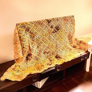 May include: A rectangular crochet throw blanket featuring an intricate openwork pattern with a variegated color palette of golden yellow, burnt orange, and deep brown tones, draped over a dark brown wooden media console with built-in speakers.