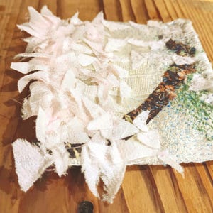 Pode incluir: Uma obra de arte têxtil quadrada com um fundo texturizado branco sujo. A obra de arte apresenta uma colagem de tiras de tecido rosa claro e branco e uma seção com um design marrom e verde. A peça é colocada em uma superfície de madeira.