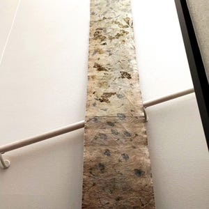May include: A long, narrow, rectangular textile runner featuring a botanical print with pressed leaves and flowers. The upper section displays light beige tones with golden-brown foliage, while the lower section transitions into a darker, earthy brown hue with blue-toned leaf imprints throughout the textured, fibrous material.