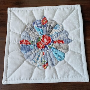 May include: Square, quilted white potholder with a central floral design. The flower is composed of blue, gray, and patterned fabric petals, with red floral accents. The white border has a quilted pattern.