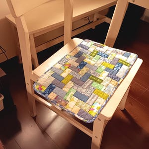 Puede incluir: Un cojín de asiento rectangular con un diseño de patchwork de aspecto tejido hecho de varios retazos de tela en tonos azul, verde, amarillo y blanco. El cojín tiene un borde estampado en azul y está colocado sobre una silla de madera blanca.
