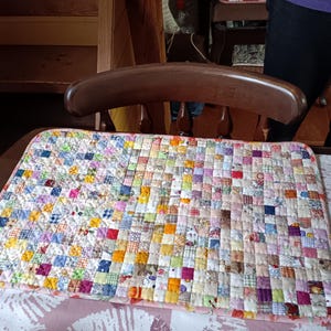 May include: A handmade patchwork quilt with a variety of colorful fabric squares. The quilt is placed on a table, with a wooden chair in the background. The quilt's design features a mix of patterns and colors.