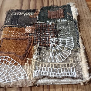 Pode incluir: Peça de arte têxtil com um design de patchwork. A composição quadrada apresenta vários tecidos texturizados em tons de castanho, preto e verde, com padrões brancos bordados em forma de teia de aranha. As bordas são desfiadas, dando-lhe um aspeto rústico.