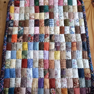 May include: A handmade, square quilted hot pad with a patchwork design. The pad features a variety of fabric squares in various colors and patterns, including floral, geometric, and solid colors. The edges are finished with a patterned border.