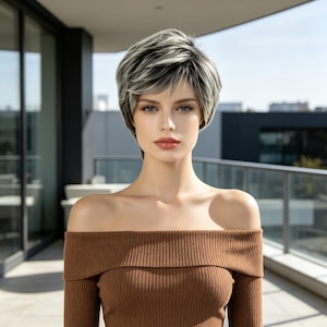 May include: A woman wearing a short, layered wig with a mix of gray and black strands. The wig has a side-swept bang and a natural-looking hairline. She is wearing a brown off-the-shoulder top. The background is a modern building with glass windows.