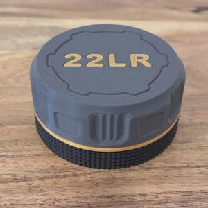 May include: A gray and black object with the text "22LR" in gold lettering. The object has a textured, ridged base and a circular top with a grooved design. The object is sitting on a wooden surface.