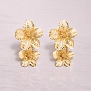Puede incluir: Un par de pendientes dorados con forma de flor. Cada pendiente presenta dos flores apiladas y detalladas con pétalos texturizados y pequeños acentos redondos en el centro. Los pendientes se muestran sobre un fondo claro.