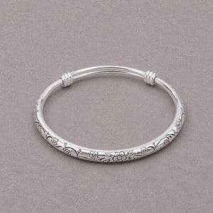May include: A silver-coloured bangle bracelet featuring an engraved floral pattern with small flowers and vines. The bracelet has a smooth, rounded band design with two distinct ribbed sections near the top, creating a continuous circular shape.