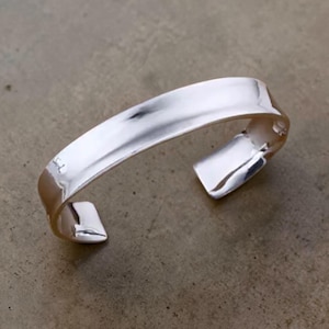 Può includere: Un bracciale a polsino in argento lucido. Il bracciale aperto presenta un design rettangolare curvo con una superficie liscia e riflettente. Il bracciale è mostrato su uno sfondo neutro.