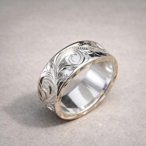 May include: A silver ring with an ornate floral pattern engraved around the band. The ring has a polished finish, reflecting light, and a smooth interior. The design features swirling leaves and delicate lines, creating a detailed and elegant appearance.