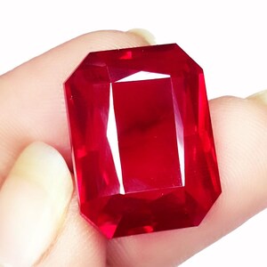 Puede incluir: Una gran gema roja vibrante, de corte rectangular. La gema tiene una superficie facetada que refleja la luz, creando reflejos brillantes. La piedra se sostiene en una mano, mostrando su tamaño y color.