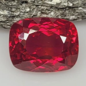 May include: A vibrant, deep red gemstone with a cushion cut, featuring numerous triangular and trapezoidal facets that create a complex, reflective surface. The stone displays a rich, saturated crimson hue with bright light reflections.