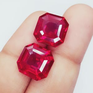 Può includere: Due gemme rosse profonde sfaccettate, taglio ottagonale. Le gemme sono tenute in una mano, mostrando le loro superfici brillanti e riflettenti. Le pietre hanno un colore ricco e saturo e sono probabilmente rubini.