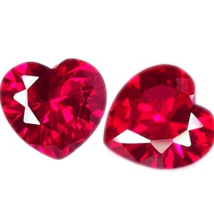 Peut inclure: Deux pierres précieuses en forme de cœur, facettées, d'une teinte rouge vif. Les gemmes sont présentées sur un fond blanc, mettant en valeur leur taille brillante et leurs surfaces réfléchissantes. Idéal pour la joaillerie ou la collection.