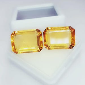 Może przedstawiać: Dwa prostokątne, fasetowane kamienie szlachetne o ciepłym żółto-pomarańczowym odcieniu. Kamienie są prezentowane w białym pudełku, ukazując ich przejrzystość i blask. Szlif kamieni podkreśla ich wewnętrzną strukturę i kolor.