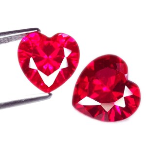 Könnte beinhalten: Zwei herzförmige, facettierte, leuchtend rote Edelsteine. Einer wird von einer silbernen Pinzette gehalten. Die Edelsteine haben einen Brillantschliff mit weißen Facetten. Die Edelsteine haben eine leuchtende, satte rote Farbe und sind vor weißem Hintergrund.