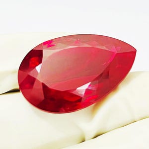 Pode incluir: Uma pedra preciosa facetada em forma de pera com uma cor vermelha profunda e vibrante. A pedra apresenta uma superfície polida com múltiplas facetas geométricas que refletem a luz, criando uma aparência brilhante na sua silhueta curva.