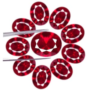 May include: A collection of nine oval-cut, deep red gemstones arranged in a circular pattern. Each stone features a faceted surface that reflects light, creating a bright white star-like pattern in the center. A pair of metal tweezers holds one stone.