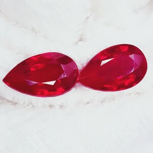 Puede incluir: Dos gemas facetadas, en forma de pera, de color rojo intenso. Las gemas se muestran sobre un fondo blanco texturizado. Las gemas tienen un alto brillo y reflejan la luz, mostrando su talla y claridad. Es probable que estas gemas sean rubíes.